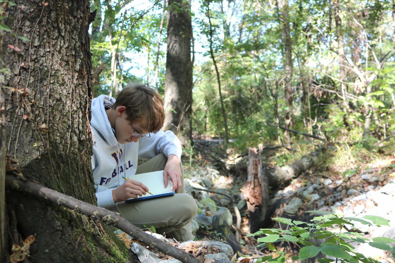 MUS student in forest writing in notebook.